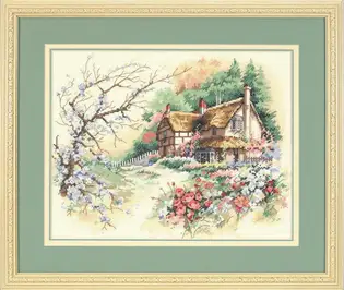 Готова вишита картина за набором Dimensions 13687 "Cottage Enchantment" (Чарівний котедж). Затишний старовинний будиночок із солом'яним дахом, розлоге весняне дерево з блакитними квітами та яскрава клумба на передньому плані, вишиті технікою лічильного хреста на світлій канві Aida 14 в оформленні із зеленим паспарту