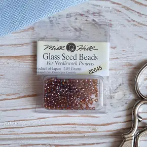 Упаковка круглого скляного бісеру Mill Hill Glass Seed Beads 11/0 (арт. 02045) цегляно-червоного кольору з райдужним напиленням. Фірмова капсула на фоні світлої стільниці поруч із канвою