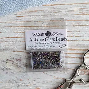 Упаковка оригінального бісеру Mill Hill Antique Glass Beads 11/0 (арт. 03012) кольору Midnight. Темні матові намистини з райдужним металевим переливом у прозорій капсулі на світлому фоні