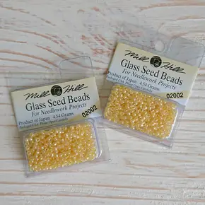 Дві упаковки оригінального круглого бісеру Mill Hill Glass Seed Beads 11/0 (арт. 02002) ніжного кремово-жовтого кольору з райдужним покриттям. Прозорі капсули з бісером на світлому дерев’яному фоні