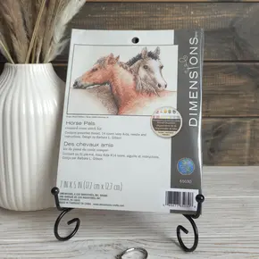 Оригінальний набір для вишивання хрестиком Dimensions 65030 Horse Pals. На упаковці зображено двох милих конячок на фоні канви кольору слонової кістки. Набір стоїть на декоративній металевій підставці на світлому столі