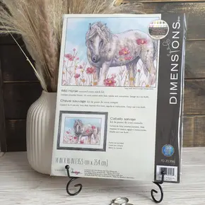 Набір для вишивання хрестиком Dimensions 70-35396 Wild Horse (Дикий кінь). На упаковці зображено величного коня серед рожевих квітів на фоні блакитного неба. Набір стоїть на чорній декоративній підставці на дерев'яному столі поруч із білою вазою