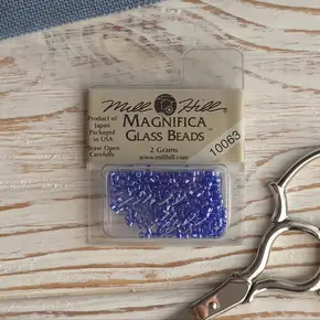 Оригінальна упаковка бісеру Mill Hill Magnifica 10063 кольору Deep Periwinkle (Глибокий барвінок) на світлому дерев'яному фоні поруч із вінтажними ножицями — купити матеріали для рукоділля та вишивки в інтернет-магазині Crazy Cross Stitch